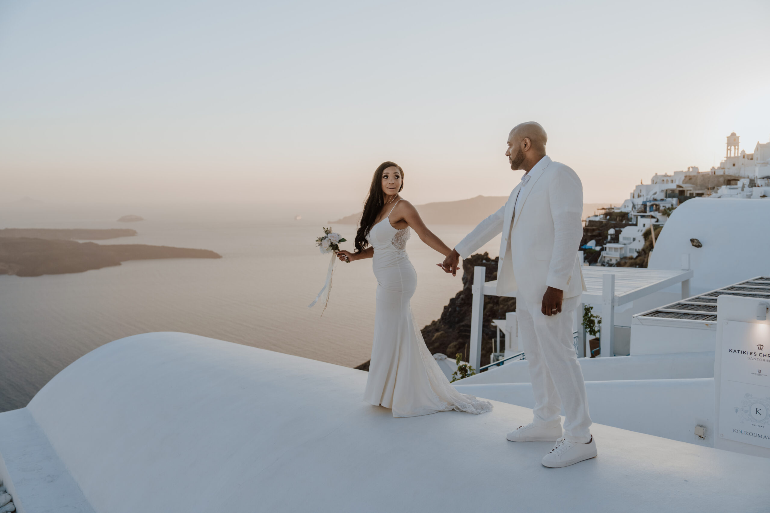 Bride and groom holding hands on a rooftop in Santorini at sunset, photographed by wedding photographer with Caldera view